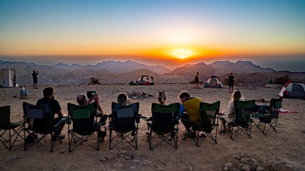 A group of hikers sitting in a row of camping chairs, looking out over a vast mountain landscape during a stunning sunset.