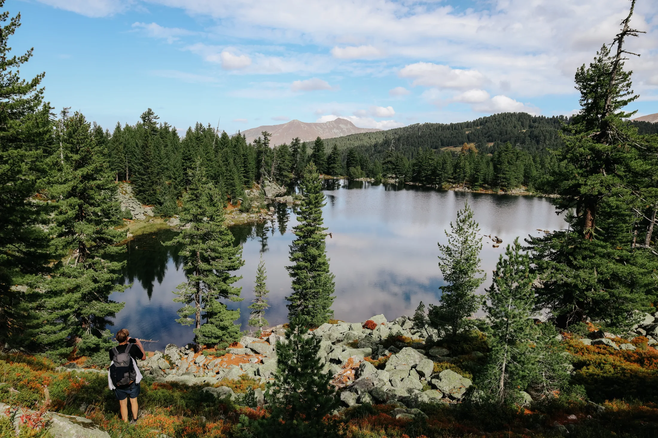 Scenic view of a small mountain lake surrounded by pine trees and distant hills under a partly cloudy sky.