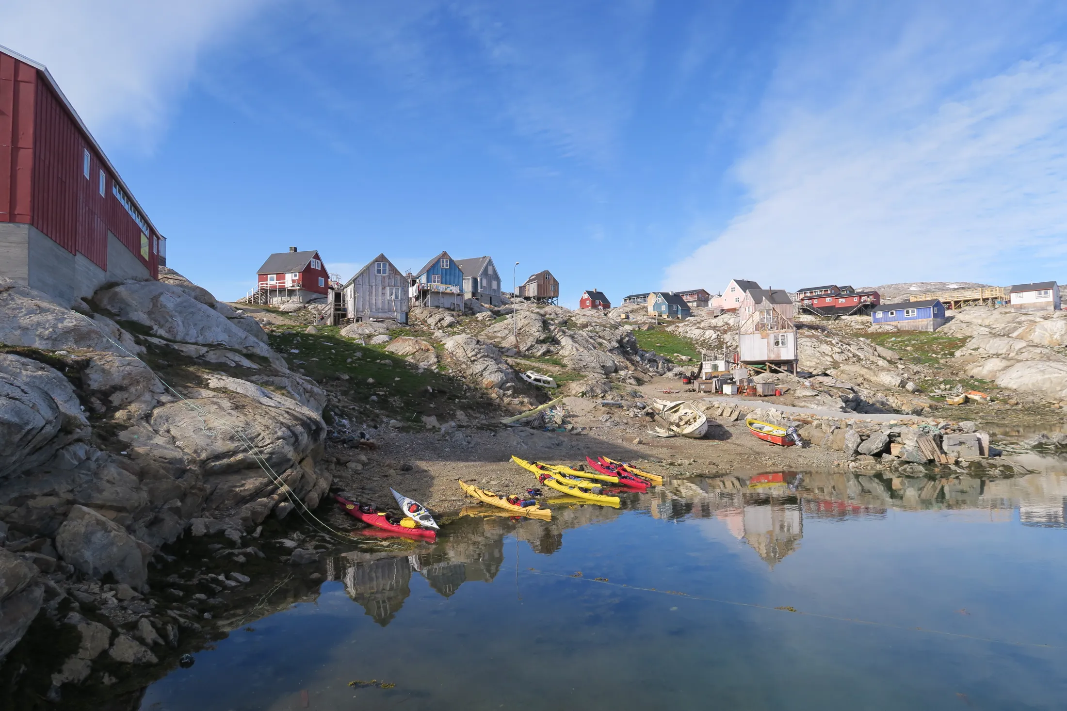 Several colorful kayaks rest on a sandy beach in a small cove in front of a Greenlandic settlement with scattered wooden houses on rocky hills under a blue sky.