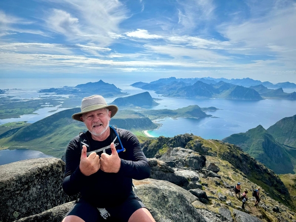 A hiker with a hat gives two thumbs up at a mountain peak overlooking a vast landscape of fjords and islands under a cloudy sky.
