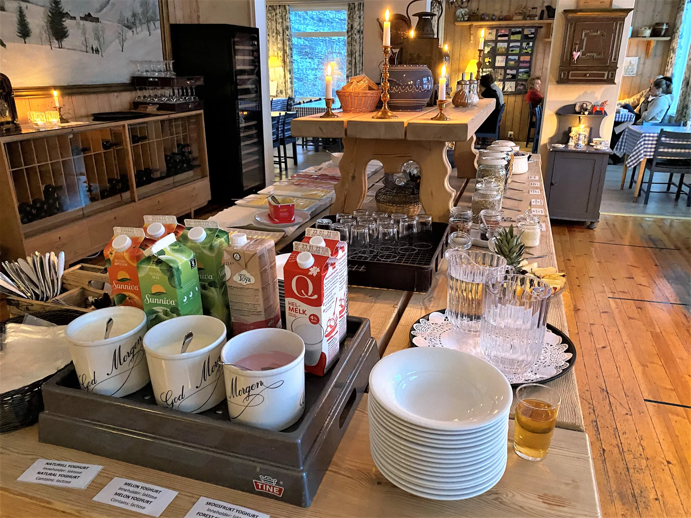 A breakfast buffet table in a rustic dining room features juice cartons, milk, bowls of yogurt, glasses, and stacked white plates.