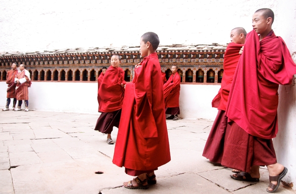 Young Buddhist monks in traditional red and maroon robes gathering in a courtyard lined with prayer wheels in Bhutan.