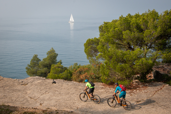 Two mountain bikers ride on a rocky path along the coast, with a sailboat on the water in the background.