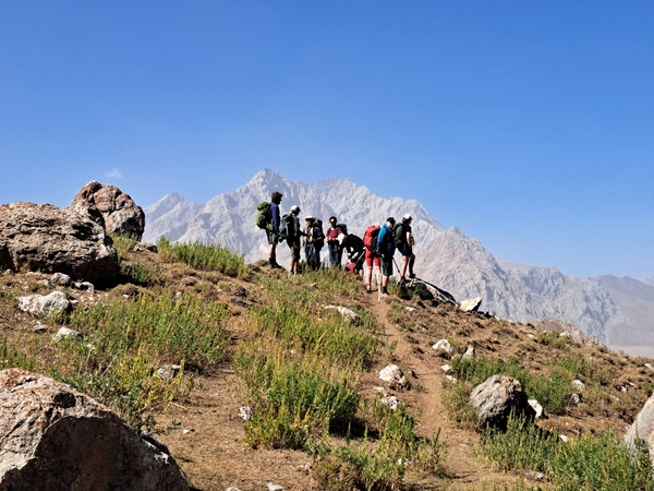 A group of hikers with backpacks stands gathered on a narrow dirt ridge trail under a clear blue sky, with a hazy, massive mountain range rising in the background.