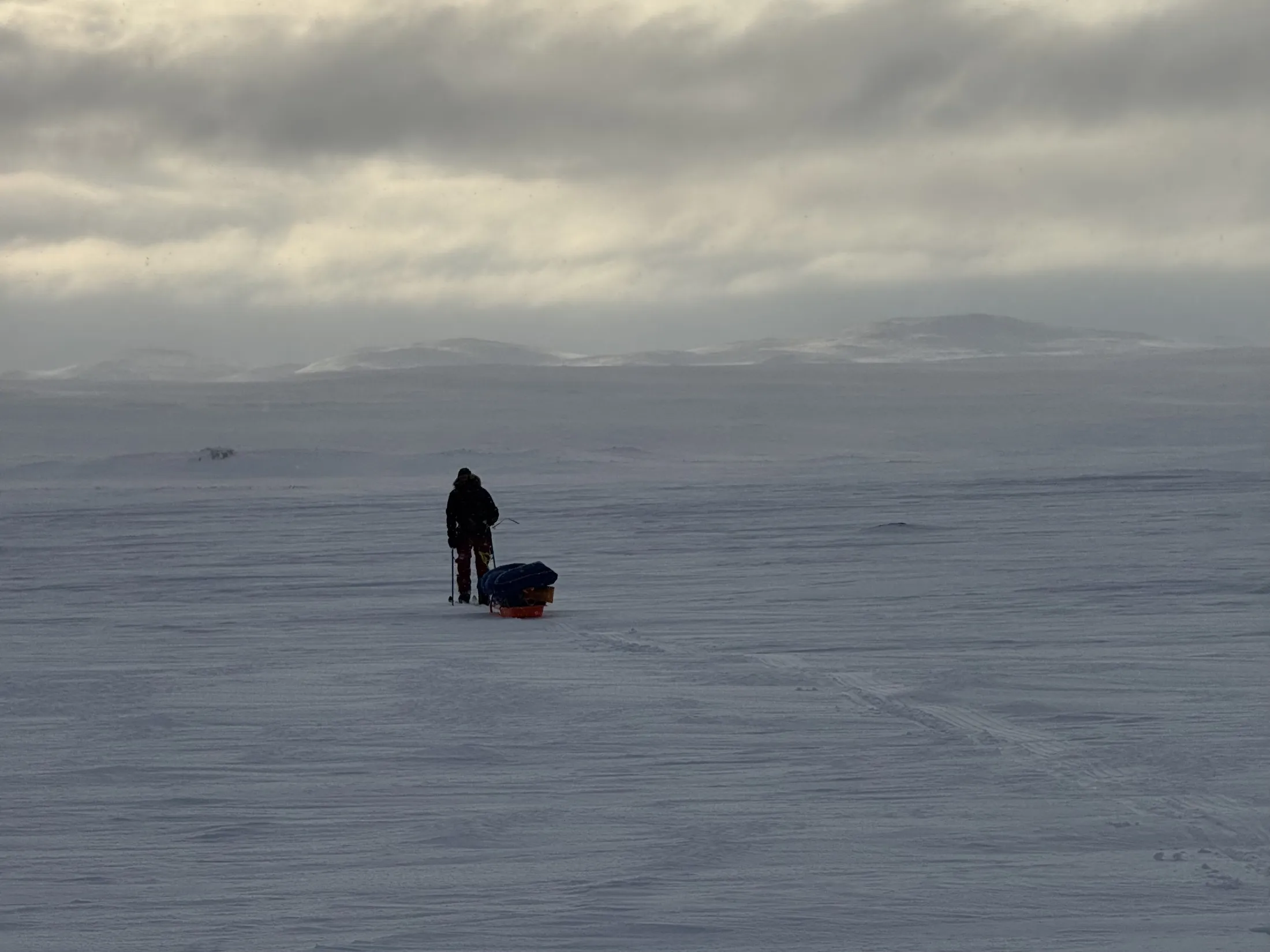 A person walking across a vast snow-covered landscape pulling a pulk, under a cloudy sky.