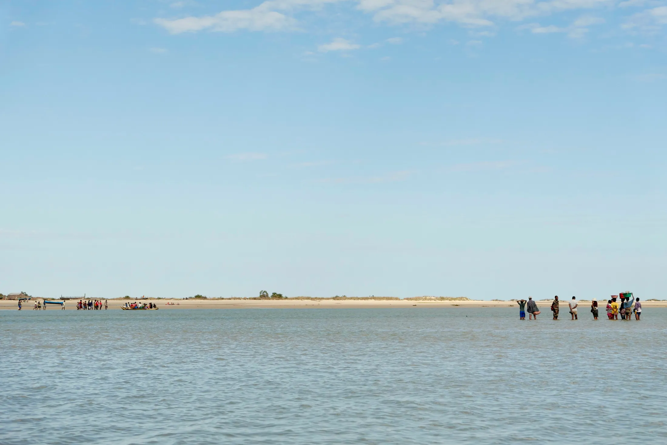 People stand in shallow water near a sandy shoreline while a small boat carrying passengers floats in the distance under a clear blue sky.