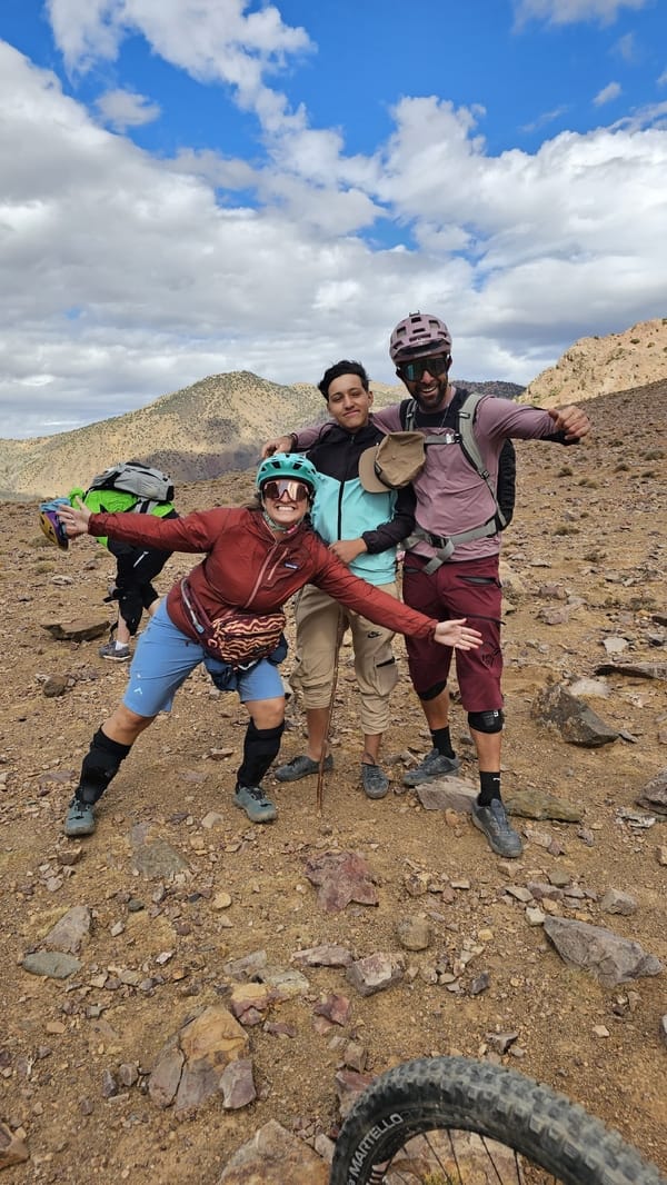 Three mountain bikers pose cheerfully in a rocky desert landscape with mountains under a cloudy sky.