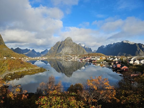 Scenic view of Reinefjorden in Lofoten, Norway, with sharp mountain peaks reflecting in the calm water and autumn foliage in the foreground.
