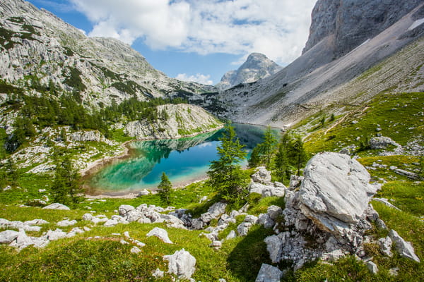 A stunning turquoise alpine lake rests in a valley surrounded by steep, rocky mountains, scattered pine trees, and grassy slopes under a partly cloudy sky.