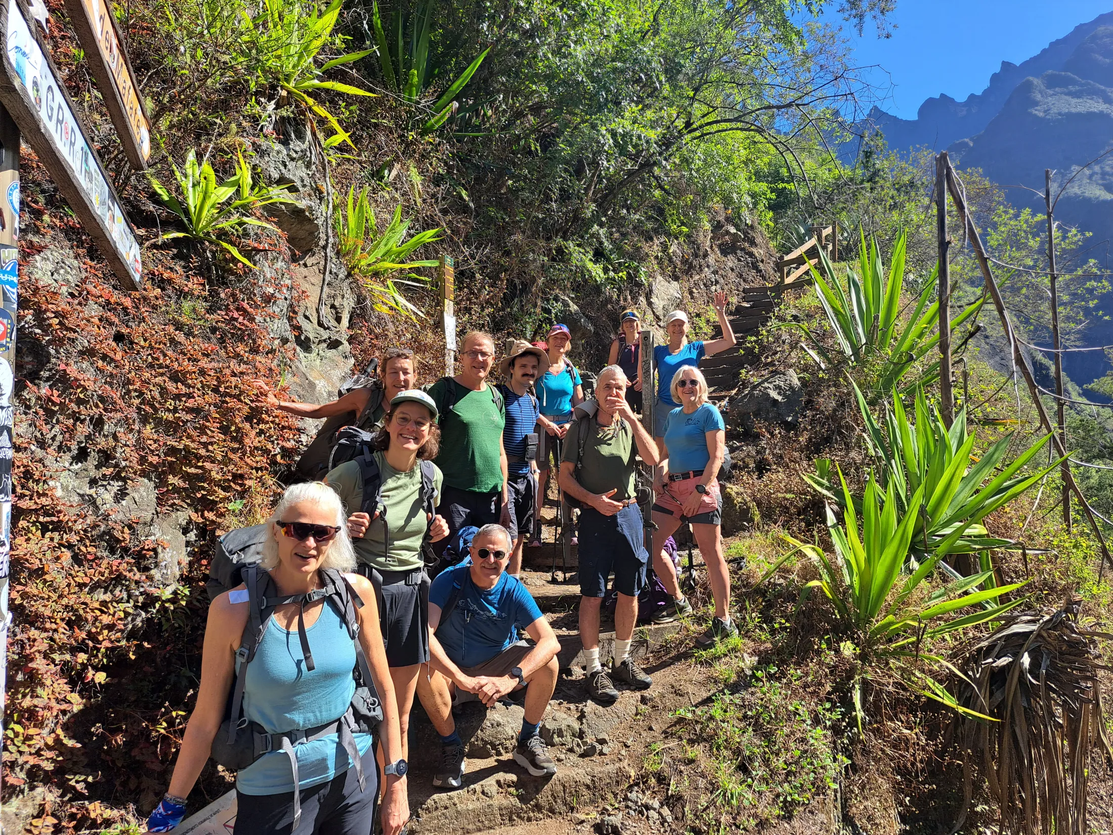 A diverse group of hikers poses happily on a steep, sunlit mountain trail surrounded by lush green tropical vegetation and agave plants.