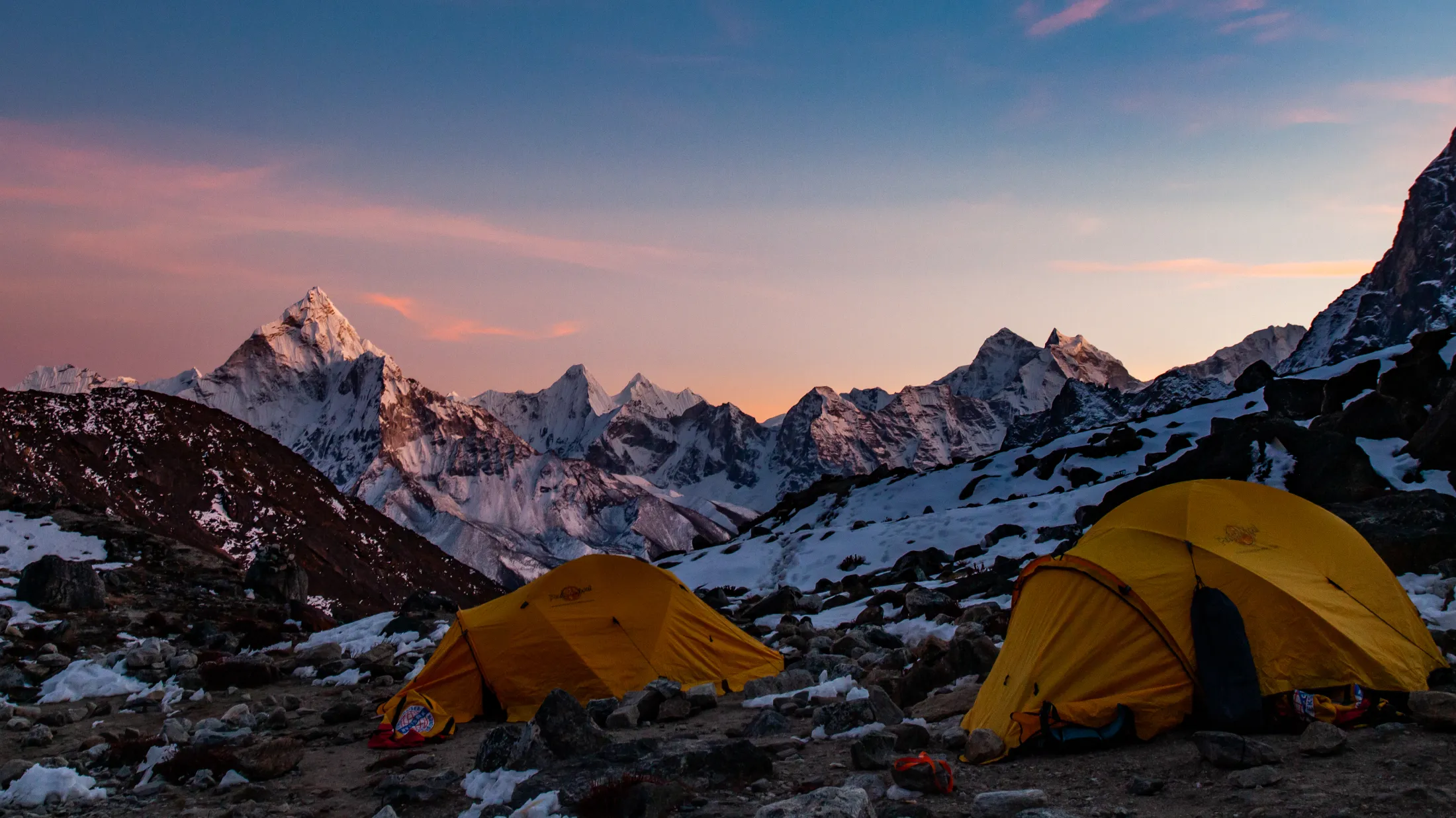 Two yellow tents sit on a rocky, snow-dusted mountainside campsite overlooking a panoramic range of rugged, snow-capped peaks illuminated by a soft pink and orange sunset sky.