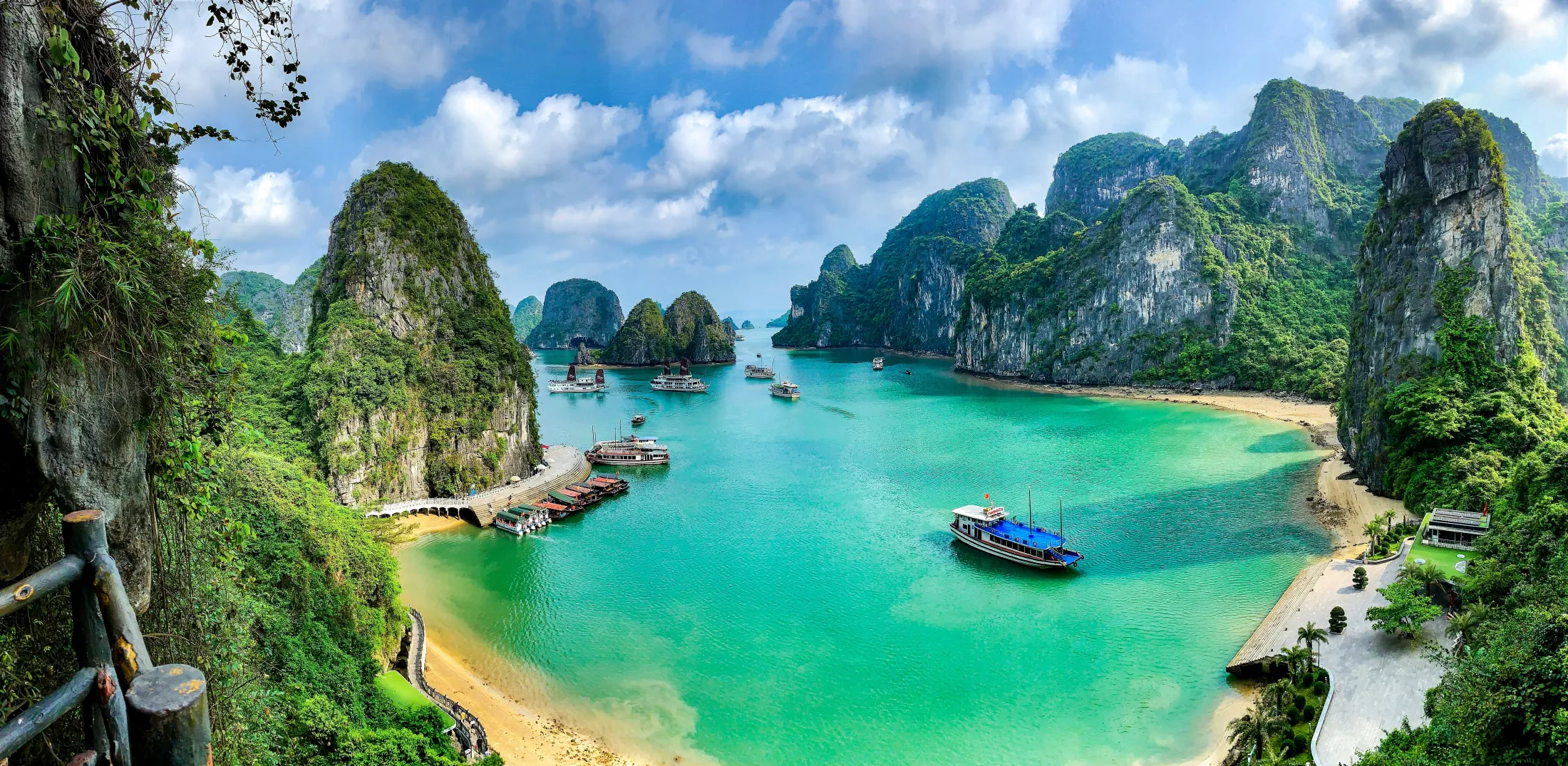 A panoramic high-angle view of Ha Long Bay in Vietnam, featuring turquoise water, sandy beaches, small boats, and dramatic limestone karsts rising from the sea under a partly cloudy sky.