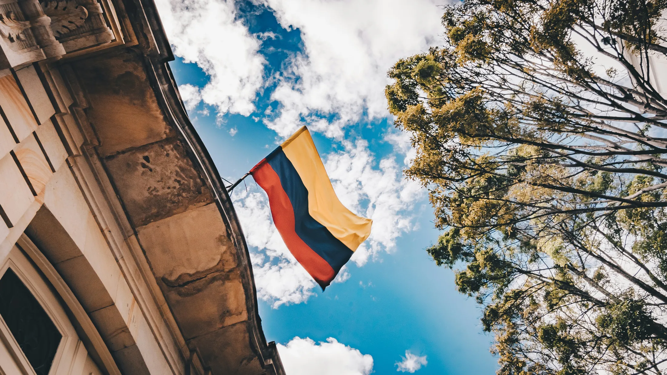 The Colombian flag waving against a cloudy blue sky, seen from below between a building and a tree.