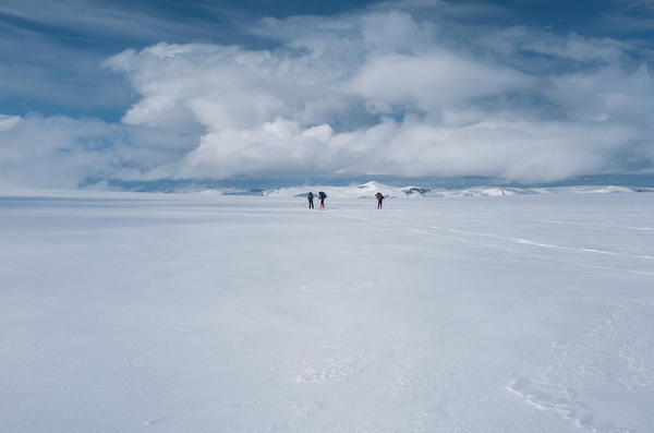 Several people cross a vast snowy landscape under a cloudy sky.