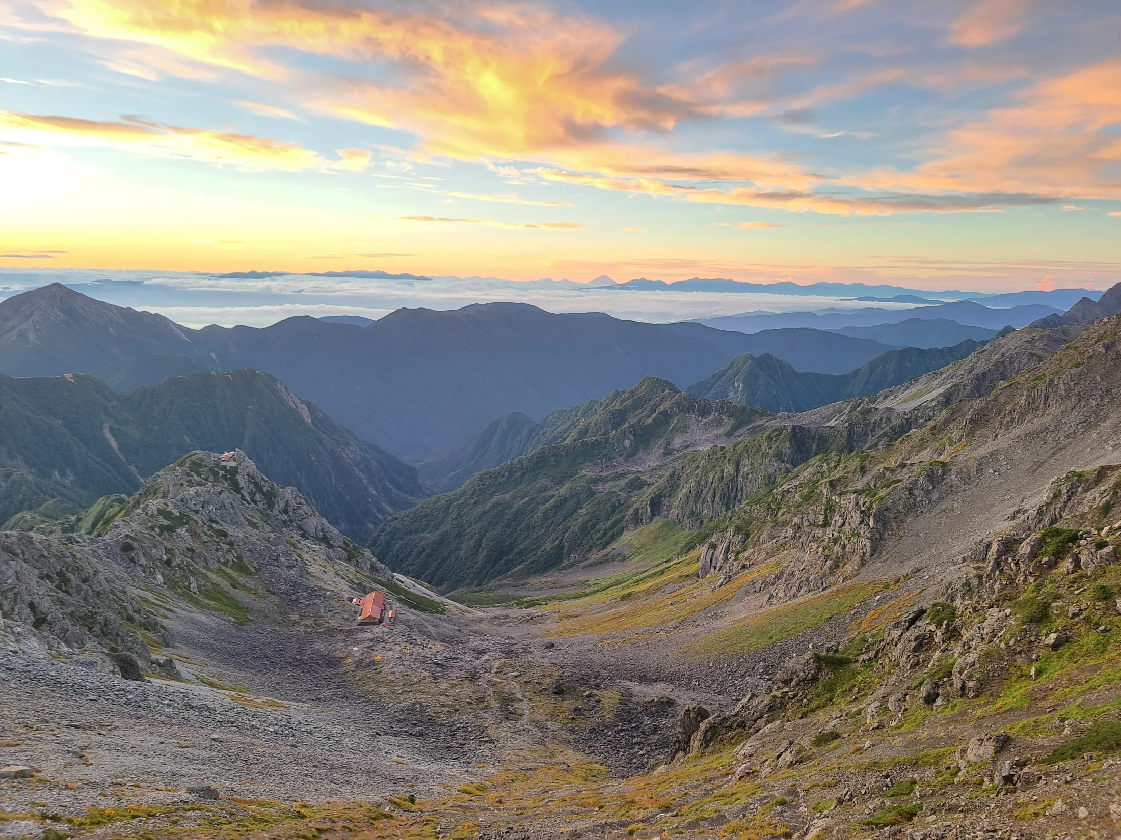 A vast alpine valley with a small red-roofed mountain hut is illuminated by a vibrant sunrise with golden and orange clouds above a sea of distant mist.