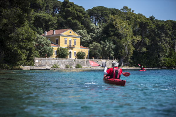 Two kayakers in red kayaks paddle across bright turquoise water toward a historic, yellow-walled villa nestled among dense green pine trees along the coast.