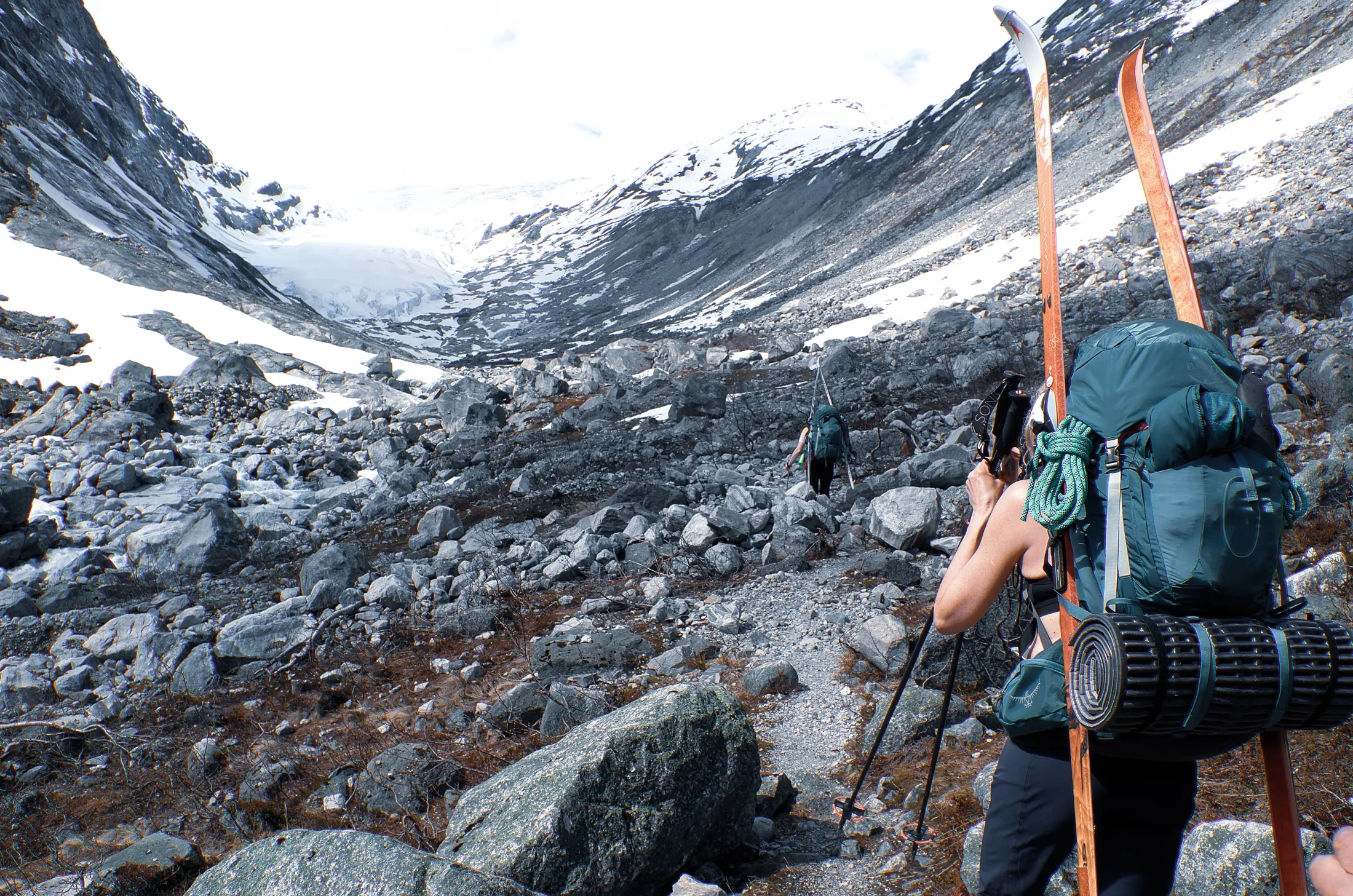 A hiker with a backpack and skis strapped to it walks through a rocky landscape with snowy mountains in the background.