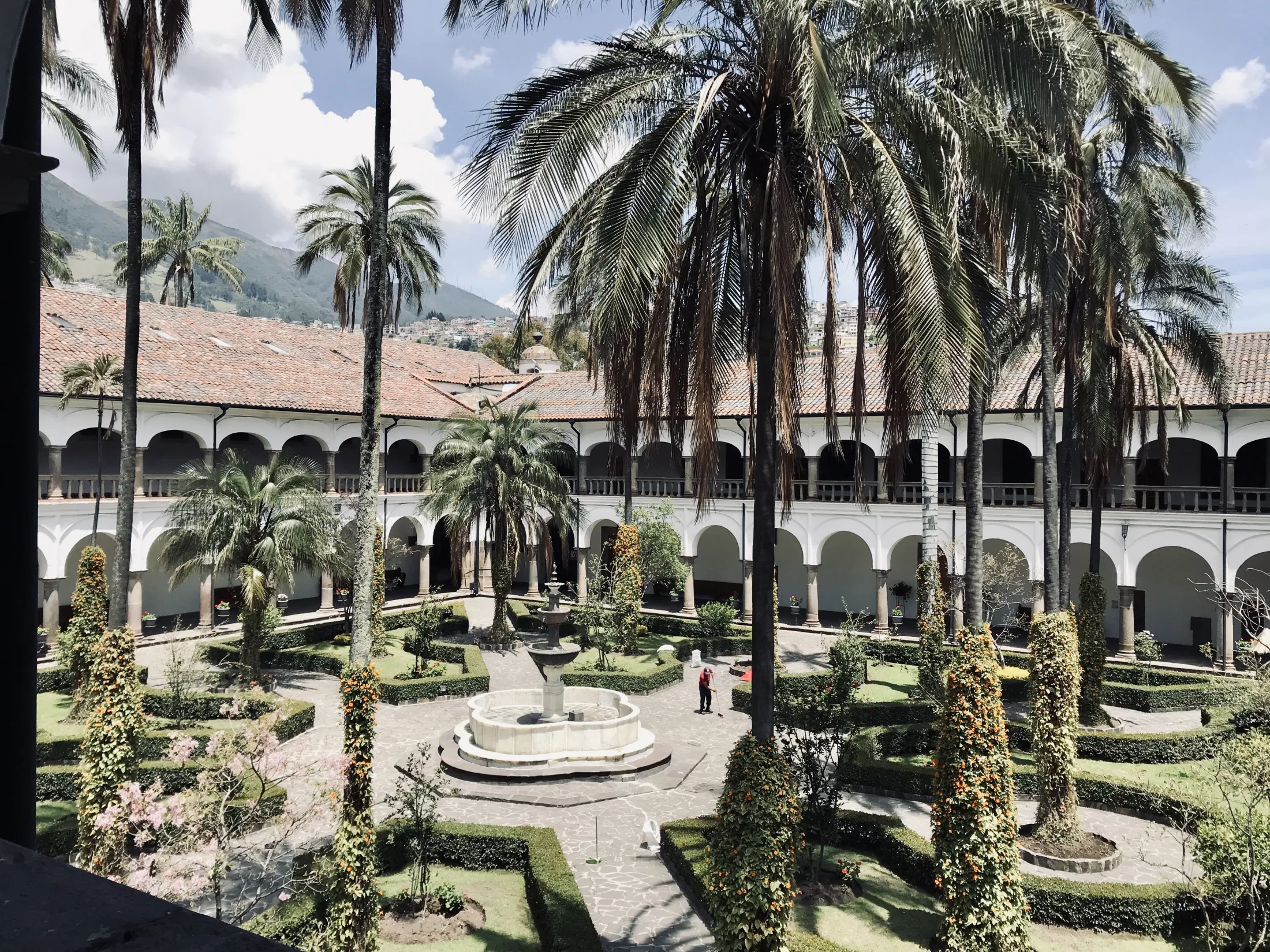 A sunlit, inner courtyard of a colonial-style building featuring a tiered stone fountain, manicured hedges, and tall palm trees surrounded by white arched corridors with terracotta tiled roofs.