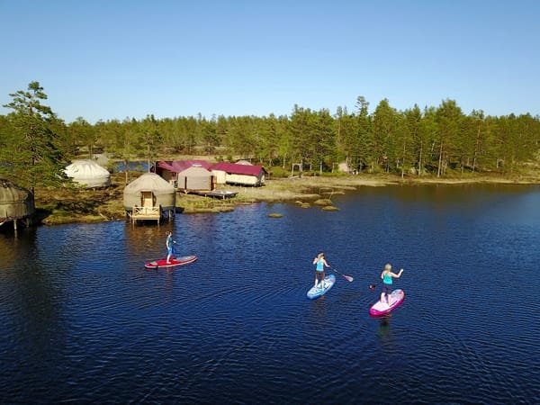 Three people paddle boarding on a calm lake near a forest shore with small cabins.