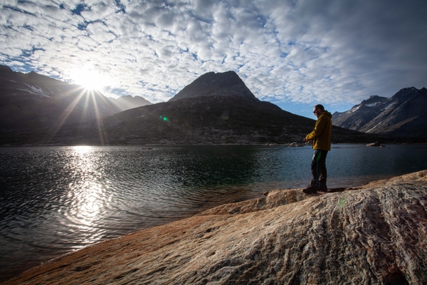 A man in a yellow jacket stands on a rocky shore fishing in a lake, while the sun bursts through the clouds over high mountains in the background.