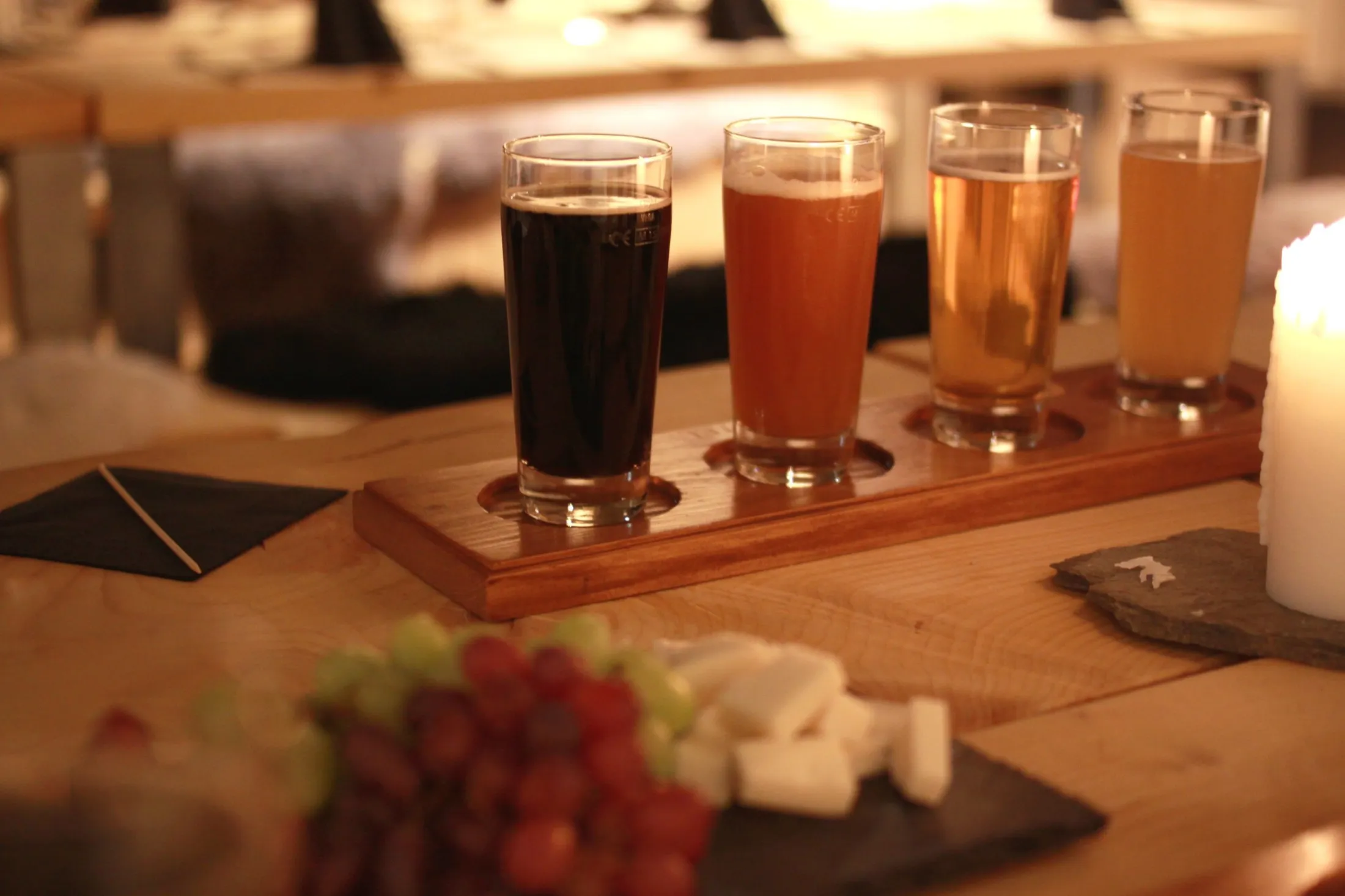A beer tasting flight of four different brews served on a wooden board next to grapes and cheese on a table illuminated by candlelight.