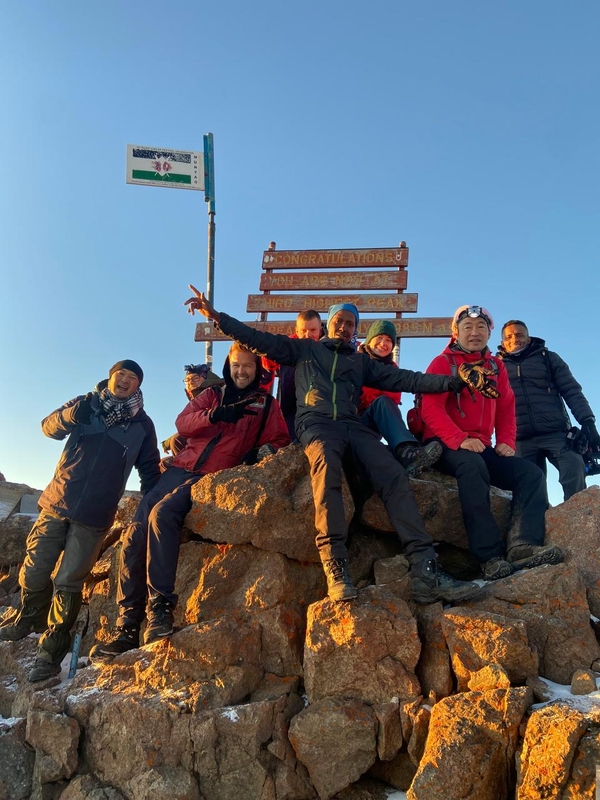 A group of happy hikers celebrates at Point Lenana summit on Mount Kenya, posing together on a rocky peak in front of a wooden congratulatory sign under a clear blue sky.