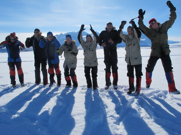 A group of eight people in winter gear stand in a snowy landscape, casting long shadows towards the camera.