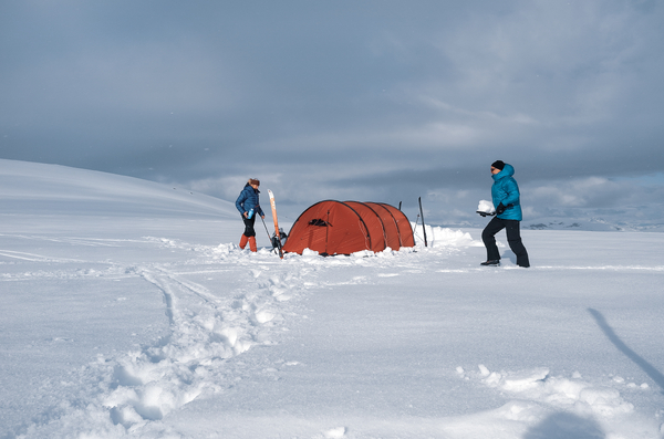 Two people camping in the snow with an orange tent under a cloudy sky.