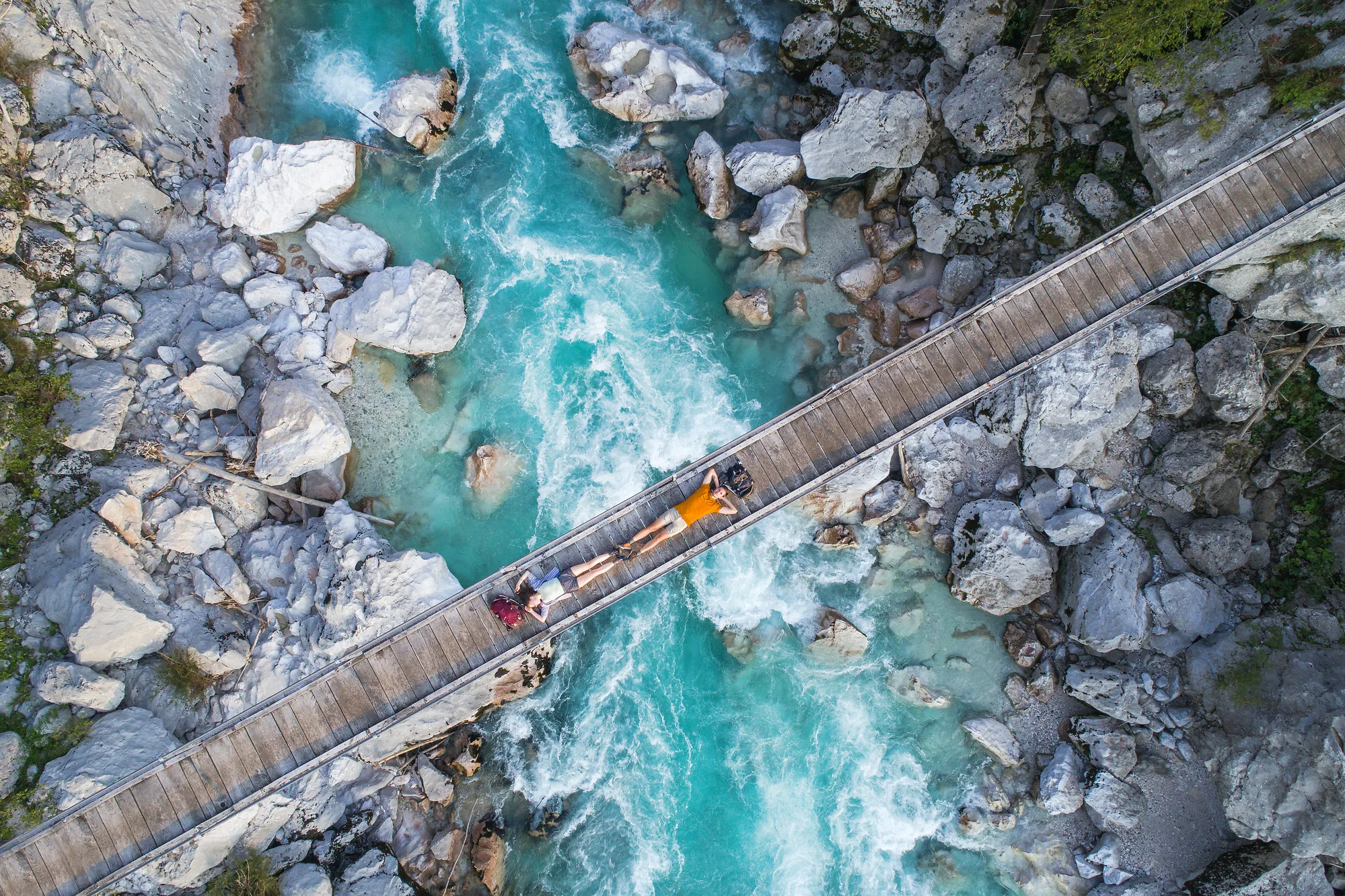 An aerial top-down view shows two hikers lying down and relaxing on a narrow wooden suspension bridge over a rushing turquoise river filled with white boulders.