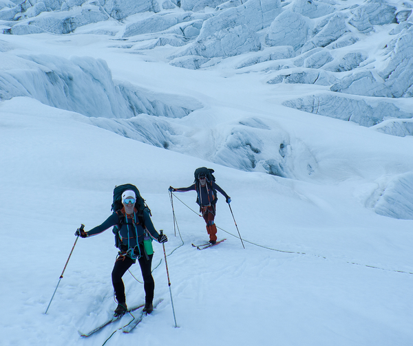 Two skiers ascending a snow-covered glacier with crevasses in the background.