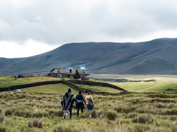 Three hikers walk across a grassy, mountainous landscape toward a distant stone house.