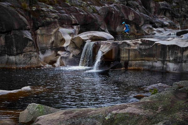 A mountain biker standing above a small waterfall flowing into a dark, calm pool surrounded by mossy rocks in a rugged, mountainous landscape.