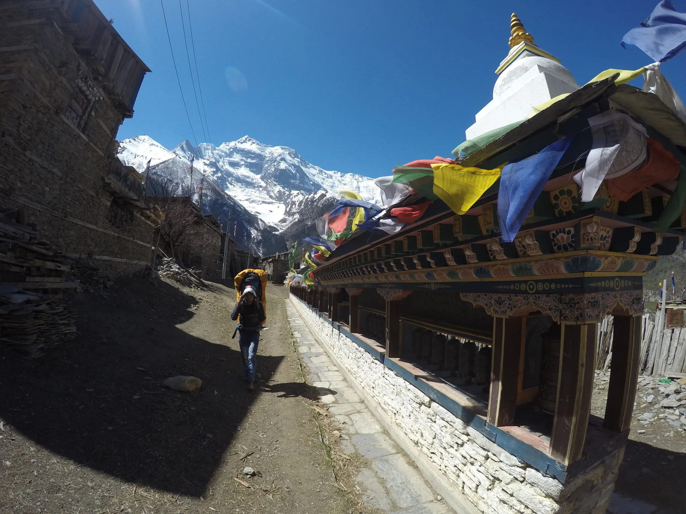 A hiker walks along a dirt path next to a long mani wall decorated with colorful prayer flags, with towering snow-covered mountains in the distance.