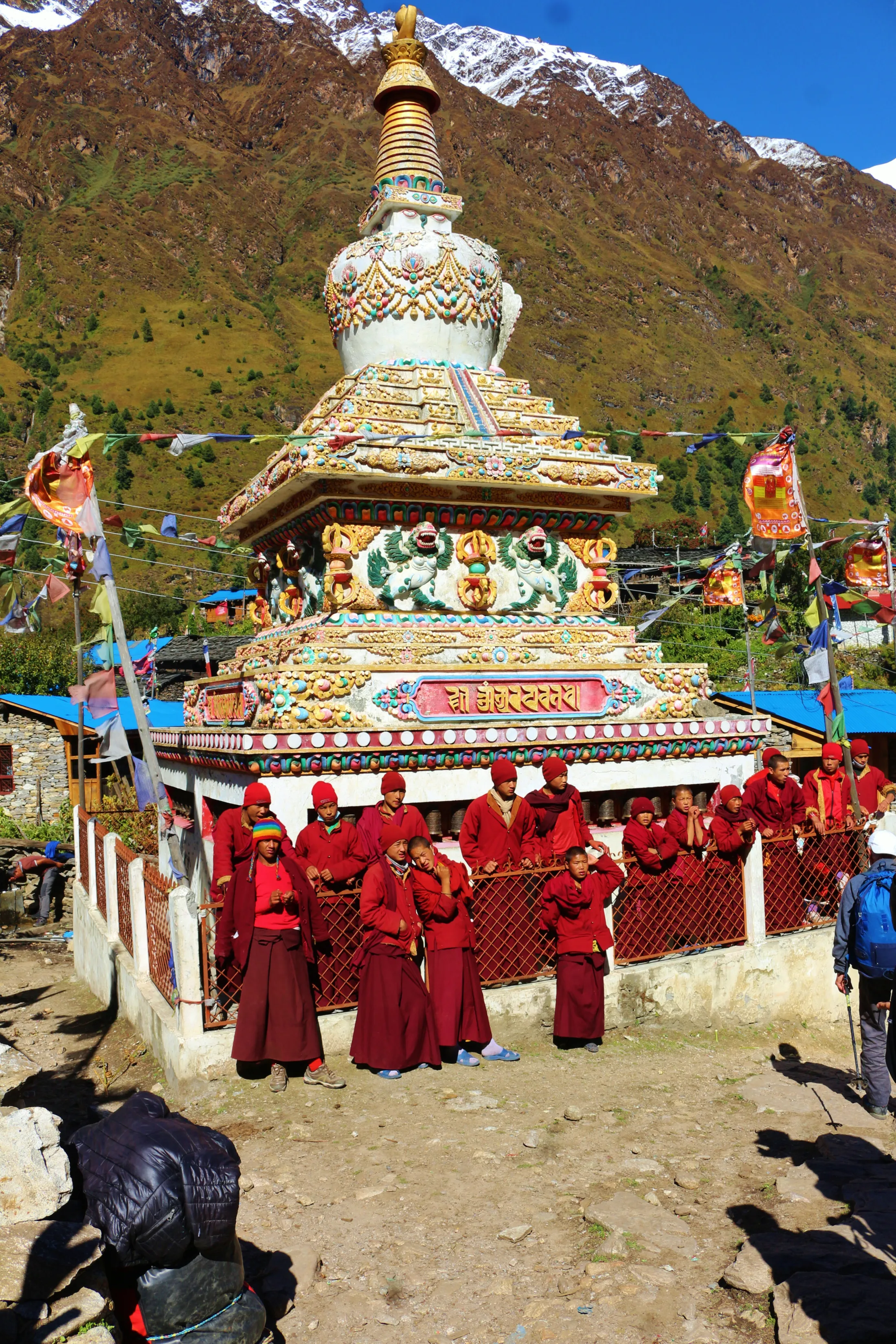 A group of Buddhist monks in red robes stands around an ornately decorated, colorful stupa at the base of a massive mountain.