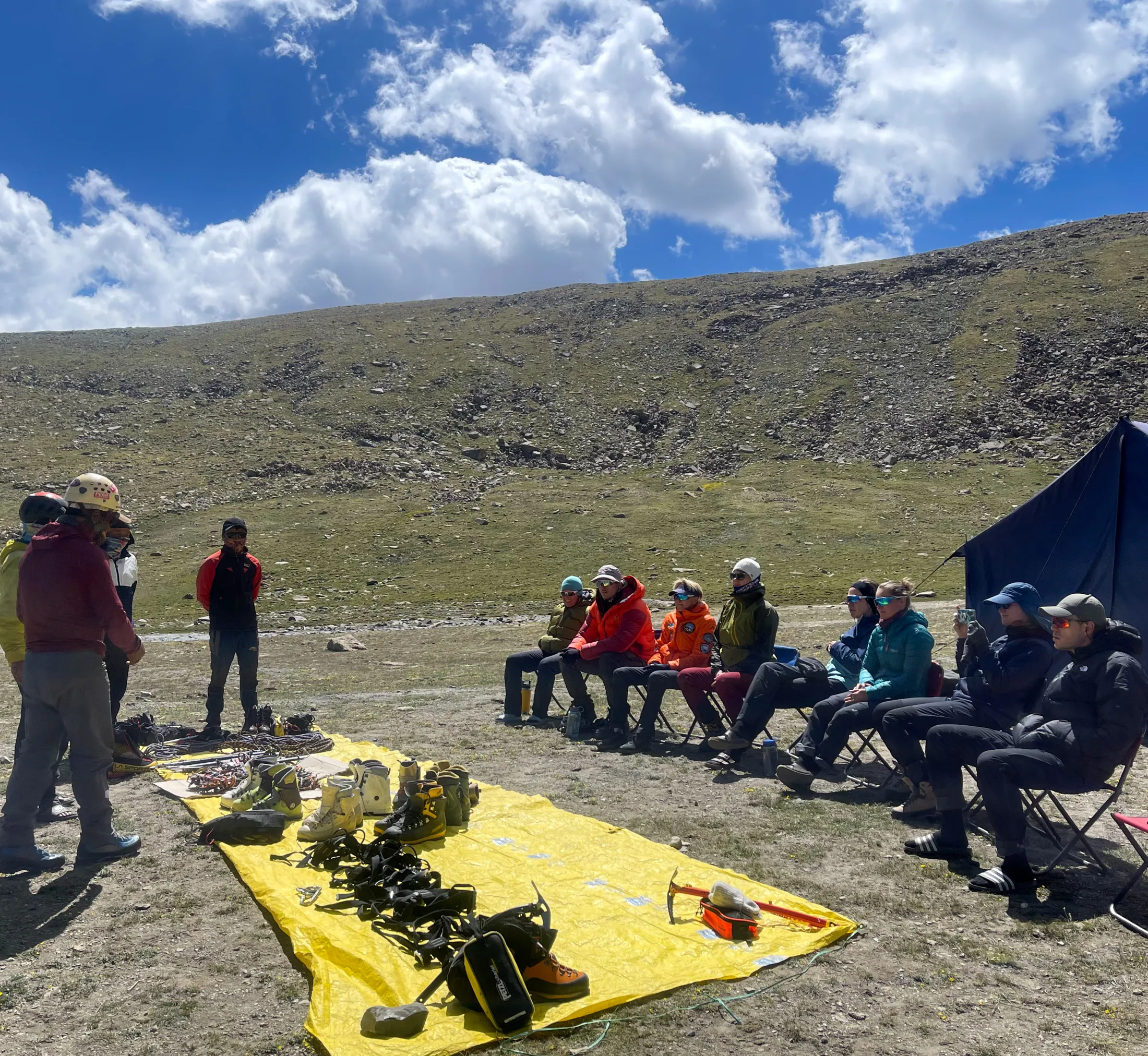 A group of mountaineers sits in chairs outdoors for a briefing, watching an instructor stand near climbing gear, boots, and harnesses laid out on a yellow tarp at a high-altitude camp under a partly cloudy sky.