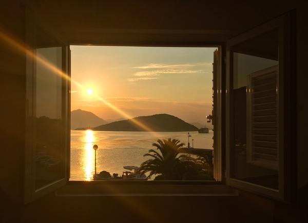 A scenic view framed by an open window looking out at a golden sunset over a calm sea, with a silhouetted island, palm trees, and a distant ferry catching the warm evening light.