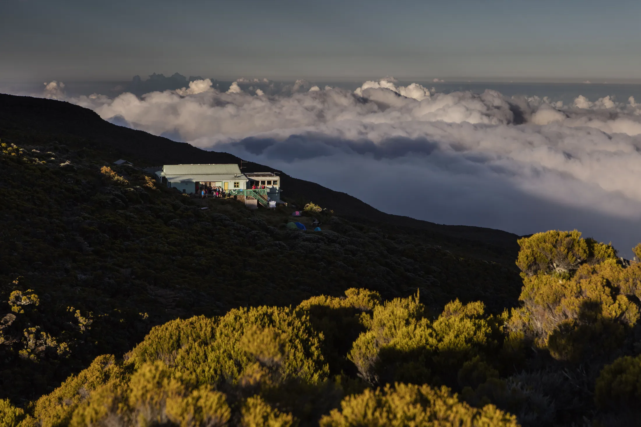 A mountain lodge sits on a dark, shrubby slope overlooking a vast sea of thick clouds under a twilight sky.