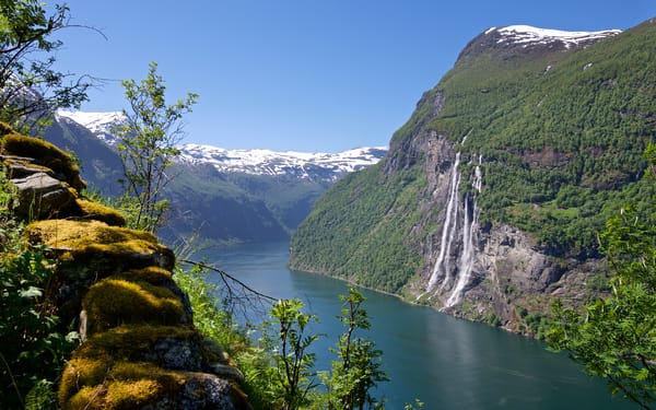 Geirangerfjord in Norway with the Seven Sisters waterfall, surrounded by green mountains and snow-capped peaks under a blue sky.