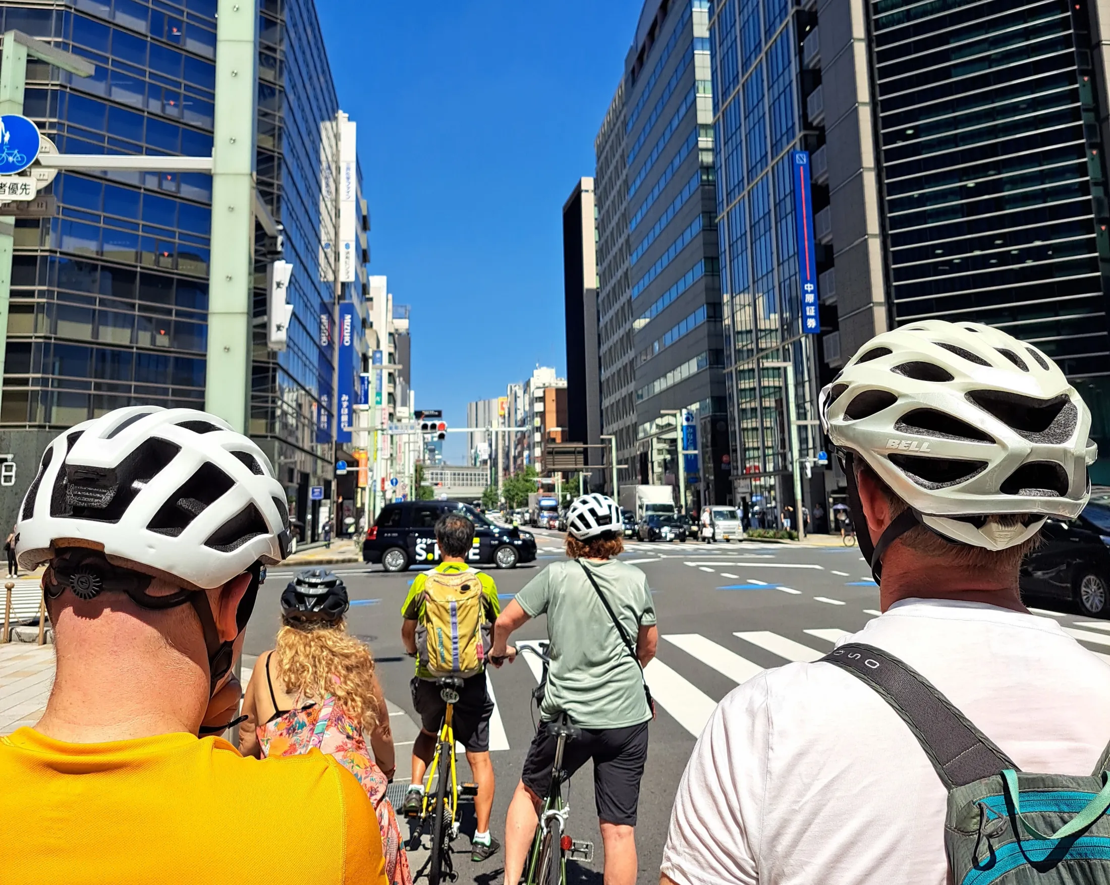 A group of cyclists wearing helmets waits on their bikes at a crosswalk in a busy city center surrounded by modern skyscrapers under a clear blue sky.