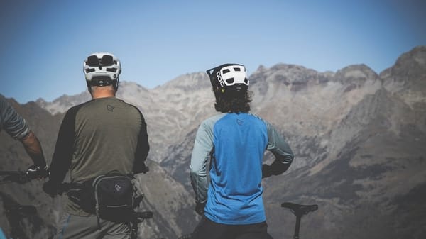 Two mountain bikers wearing helmets standing on a peak overlooking a vast mountain range under a blue sky.