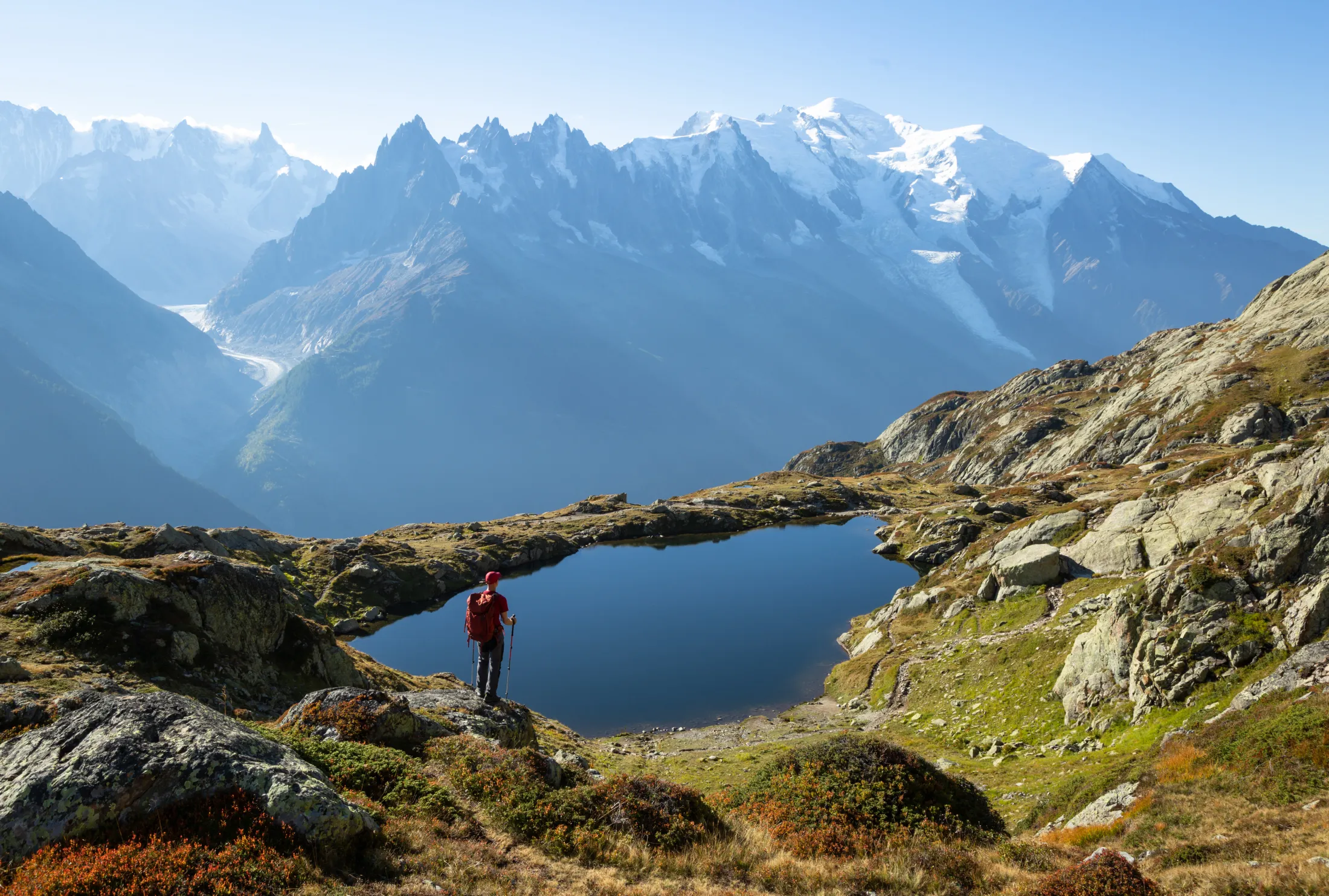 Person by a small water in the mountains looking at a larger snow covered peak in the summer.