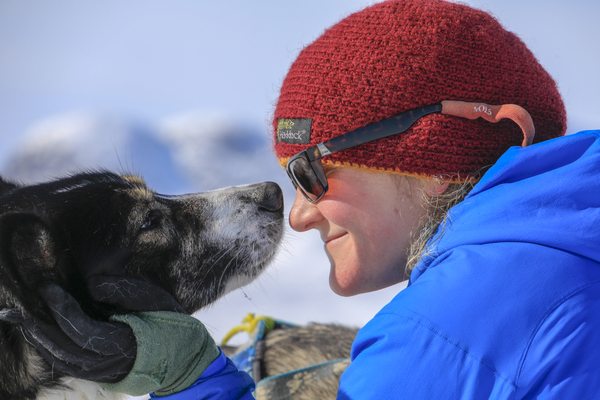 A person in a red beanie and blue jacket touches noses affectionately with a sled dog in a snowy landscape.