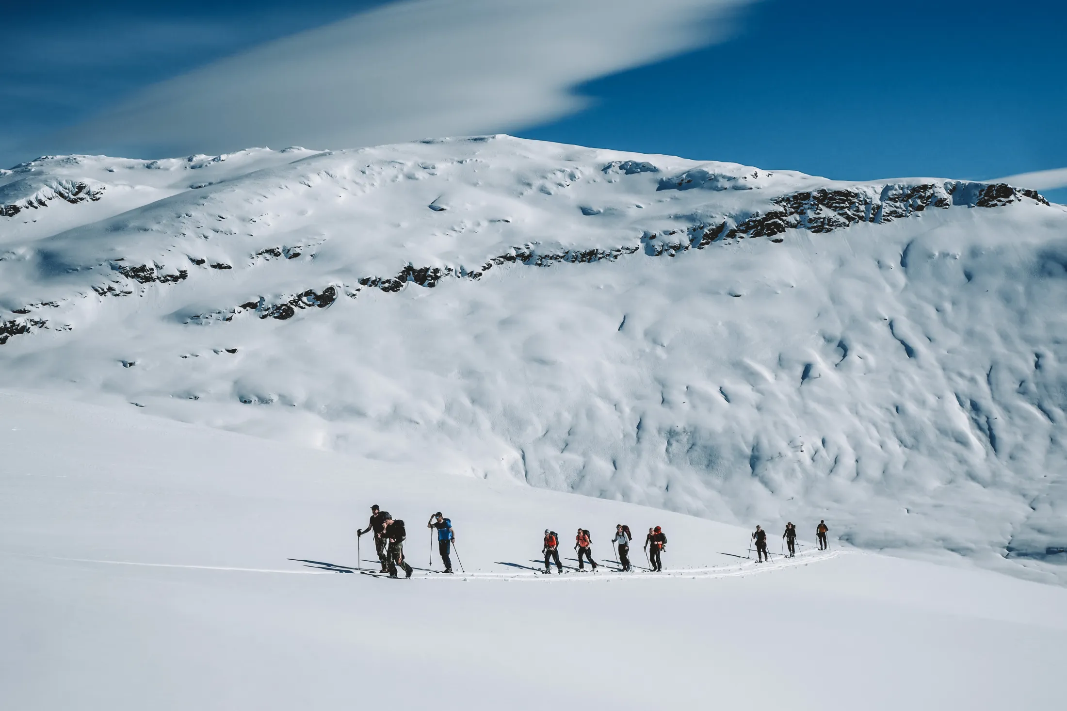 A group of people on skis travels in a long line across a snow-covered mountain landscape under a blue sky.