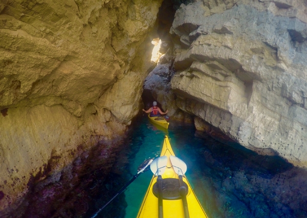 A first-person view from a yellow kayak navigating through a narrow, rocky sea cave with clear turquoise water, following another kayaker ahead toward a sunlit opening.
