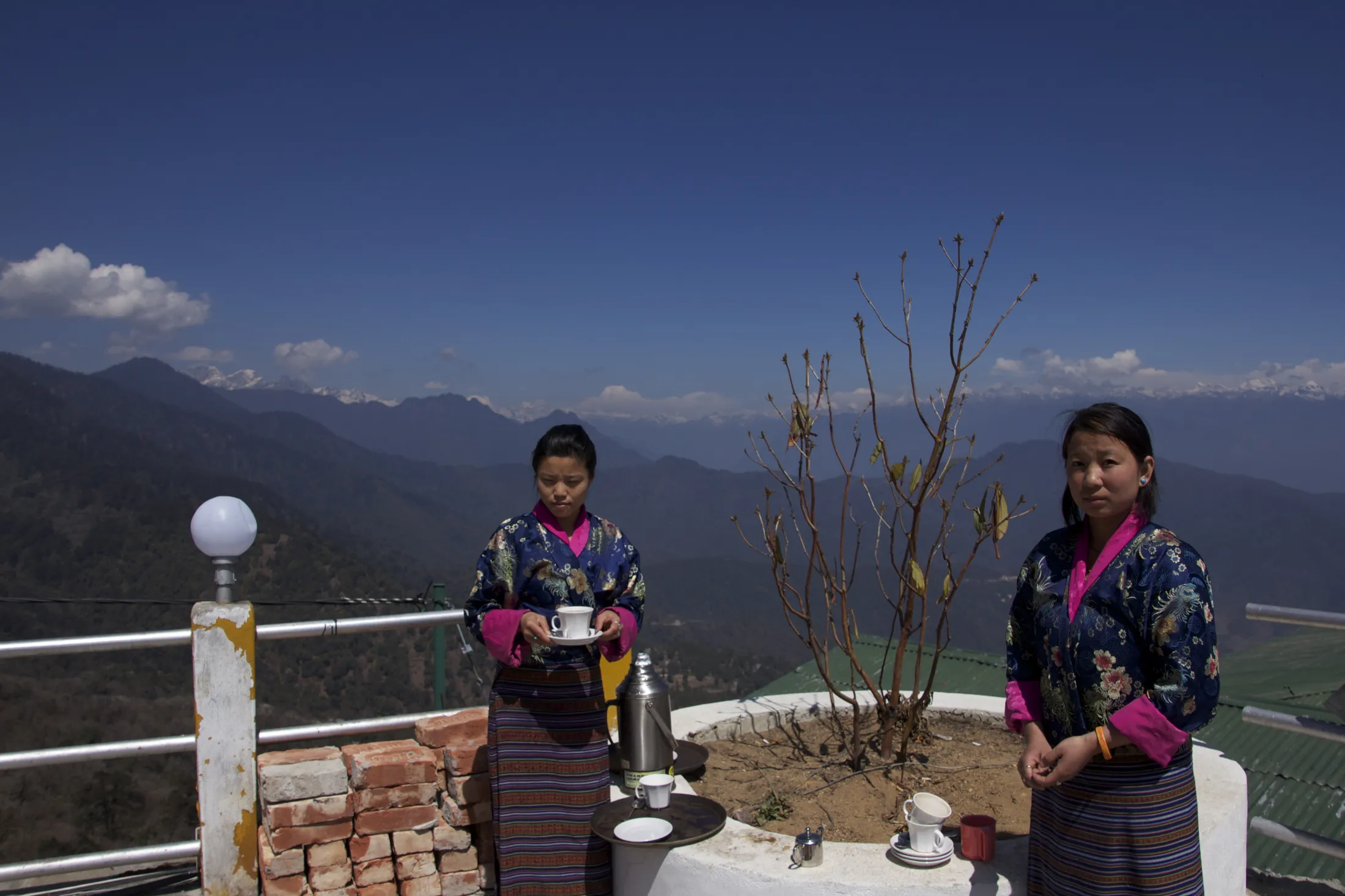 Two Bhutanese women in traditional attire serving tea outdoors on a mountain viewpoint with distant Himalayan peaks in the background.