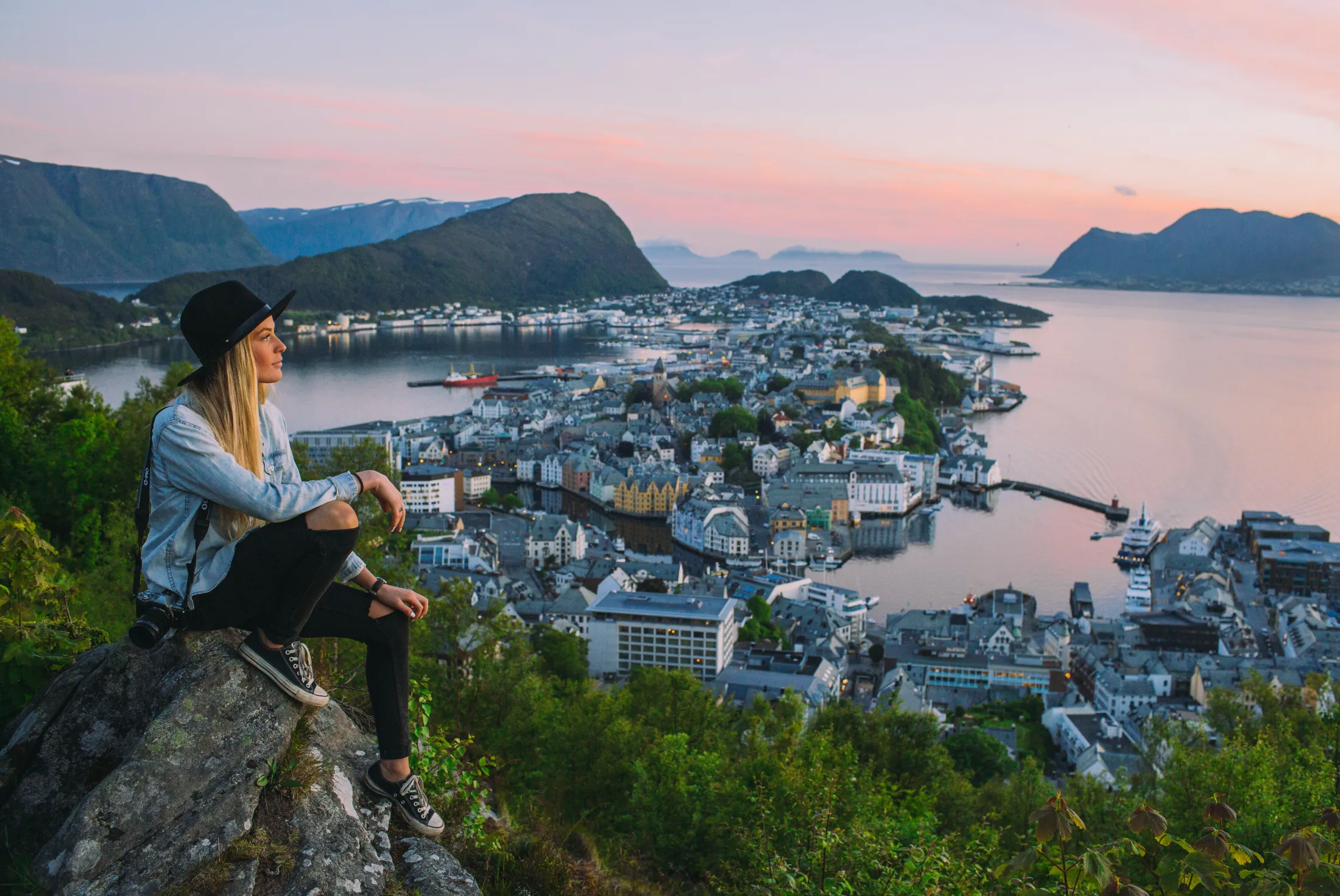 A woman sits at Aksla viewpoint, bathed in the golden light of the setting sun as dusk settles over Ålesund’s art nouveau rooftops and surrounding fjords.
