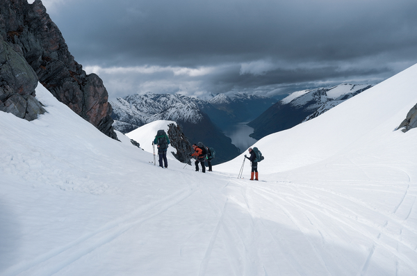 Hikers walking down a snowy mountain slope with a view of a fjord under a cloudy sky.
