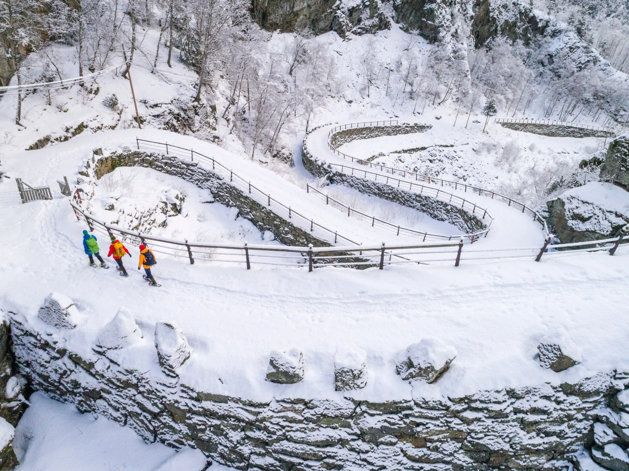 Three hikers in colorful jackets walk along a winding, snow-covered stone road cut into a steep hillside.