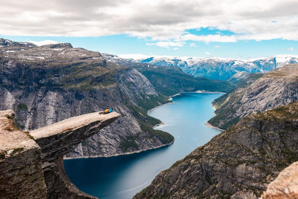 A view of Trolltunga in Norway, with the iconic rock formation protruding over a steep cliff overlooking a blue fjord surrounded by mountains.