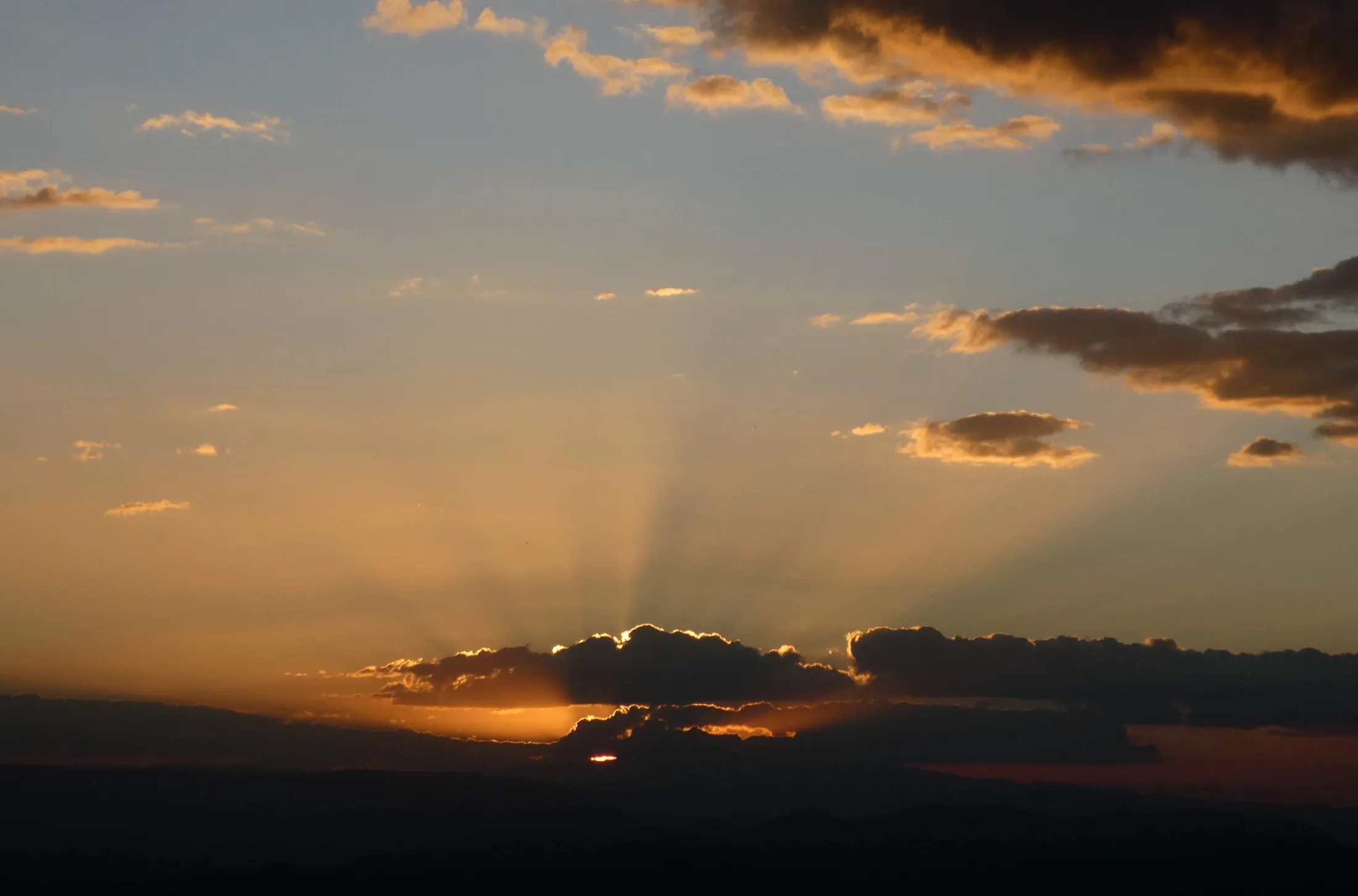 The golden rays of a setting sun break through dark clouds over a silhouetted mountain landscape, illuminating the sky with a warm, orange glow.